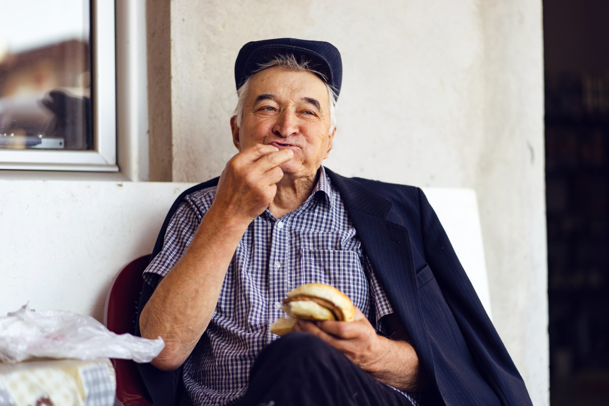 senior man eating fast food hamburger burger while sitting by the window on the balcony or entrance door at home wearing cap and jacket over his shoulders